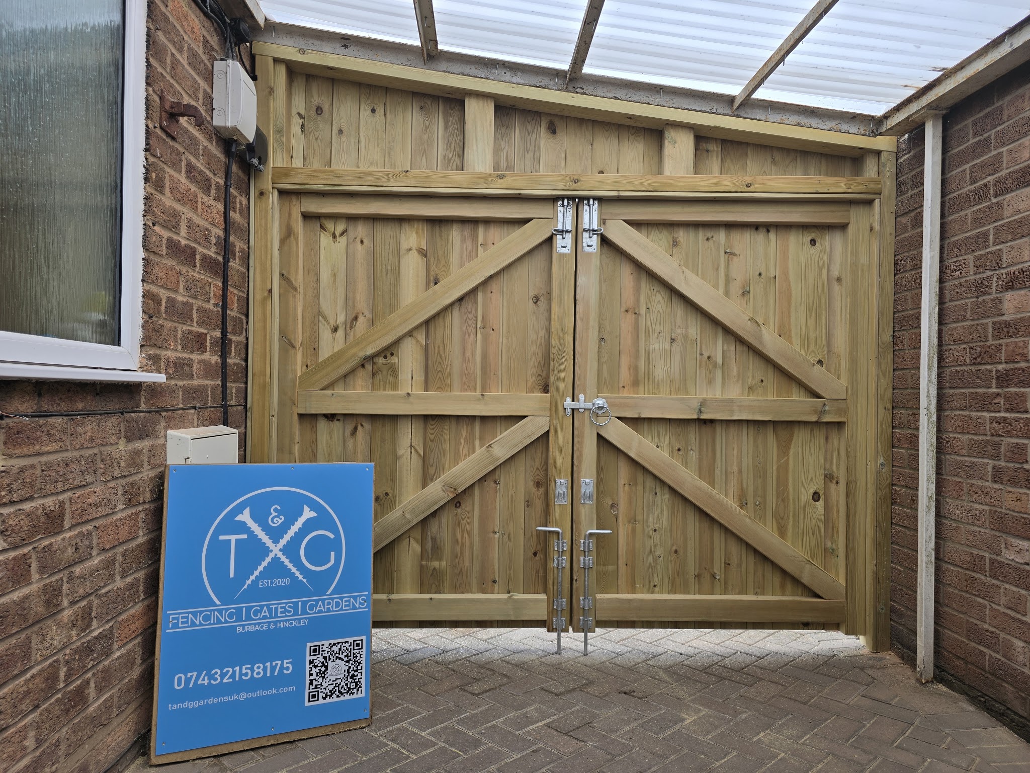 Ledged and braced natural timber double gates under a clear-roofed side canopy, with the T&G sign board