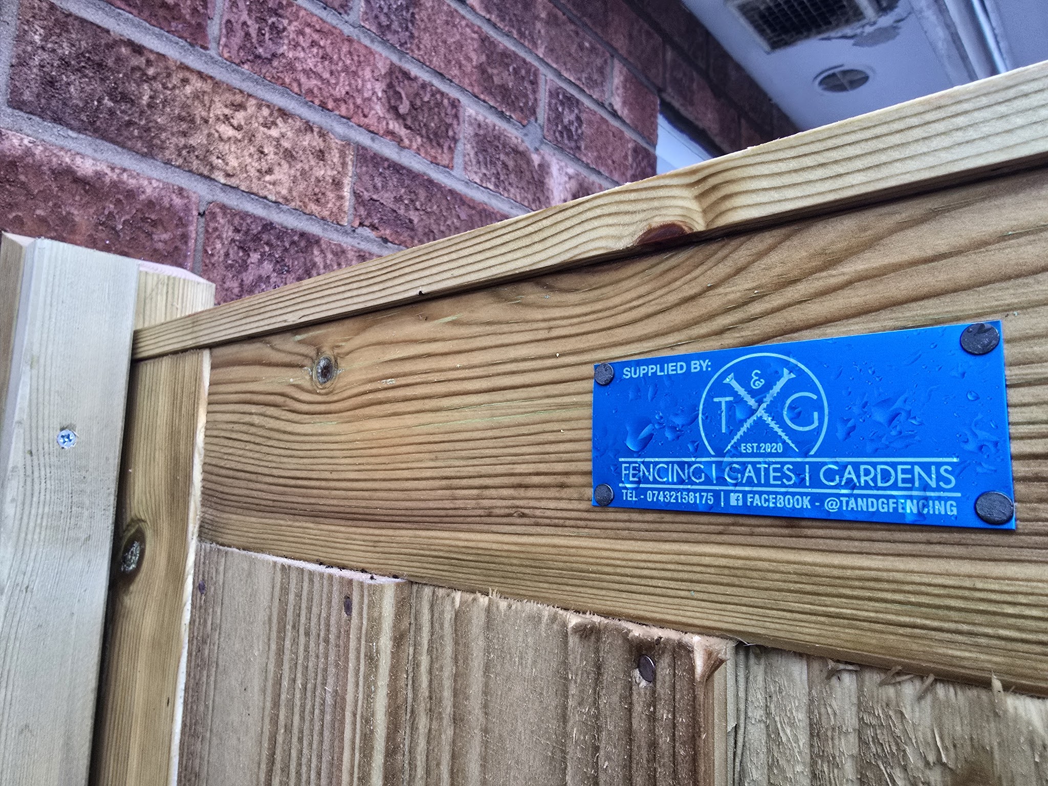 Blue ‘Supplied by T&G’ plate screwed to the top rail of a new fence, with the phone number and Facebook name
