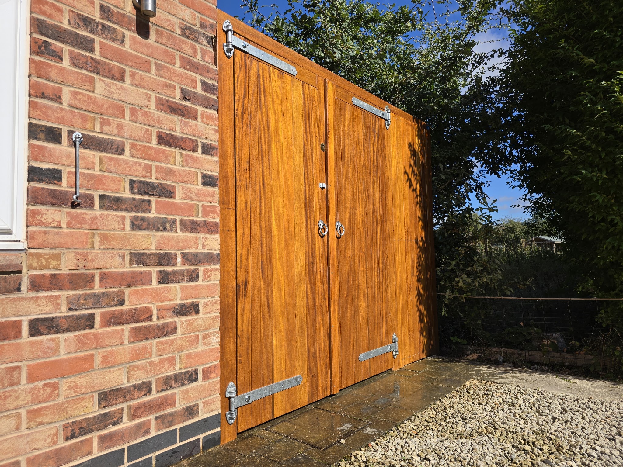 Tall double timber gates stained a rich golden colour, hung on black strap hinges beside a brick house