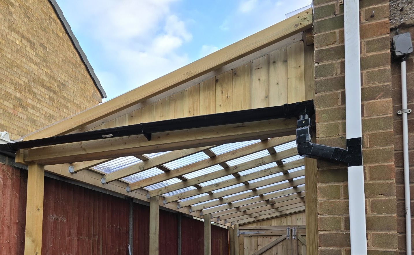 Timber canopy frame with a boarded gable, clear roof sheets on timber rafters and a ledged and braced gate at the far end