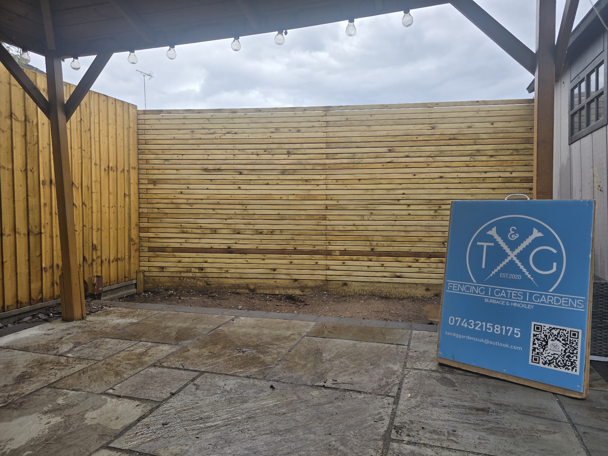 Horizontal slatted timber fence under a timber pergola strung with festoon lights, with the T&G sign board
