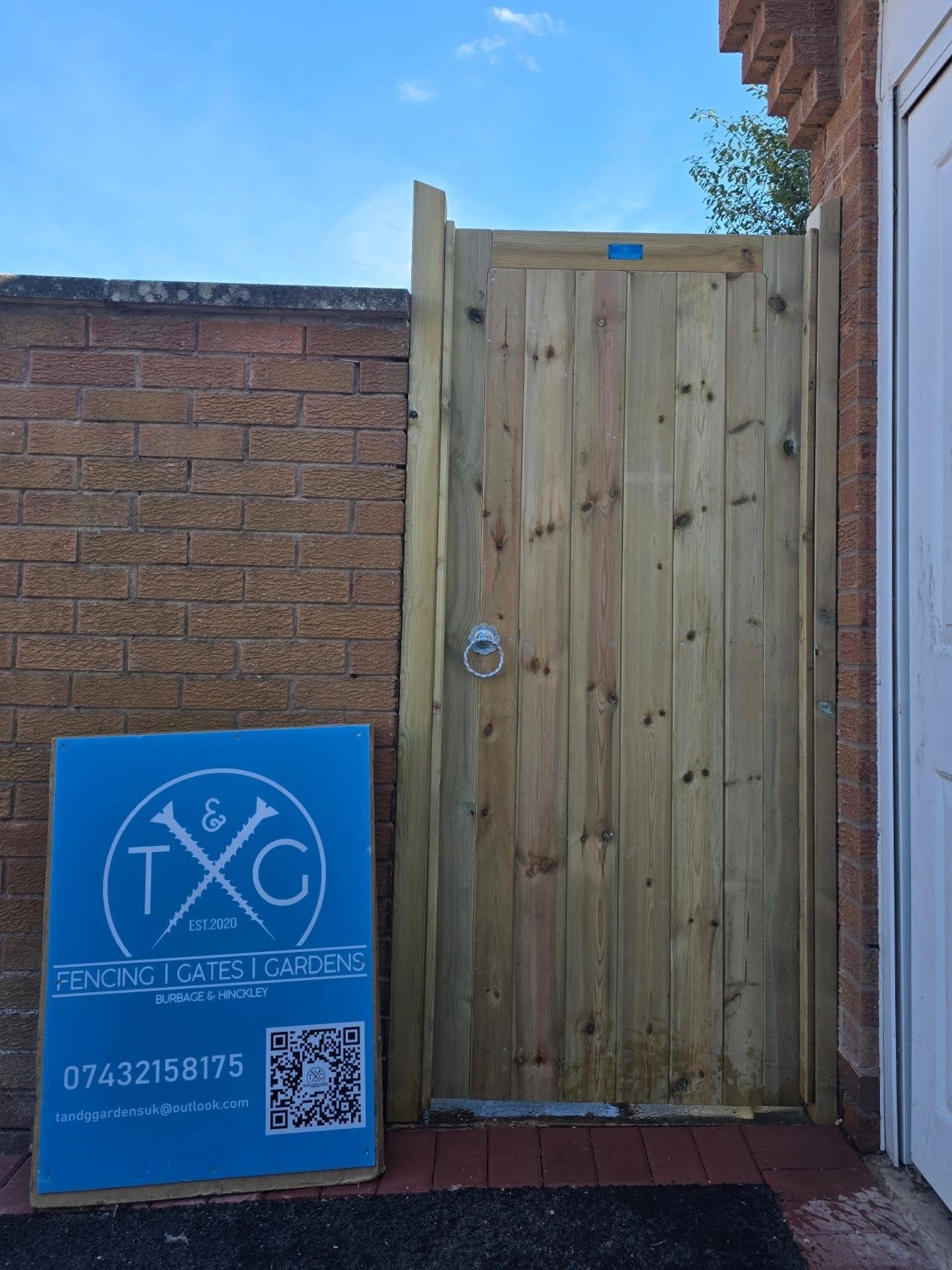 Tall natural timber side gate fitted between a brick wall and a house, with the T&G sign board in front