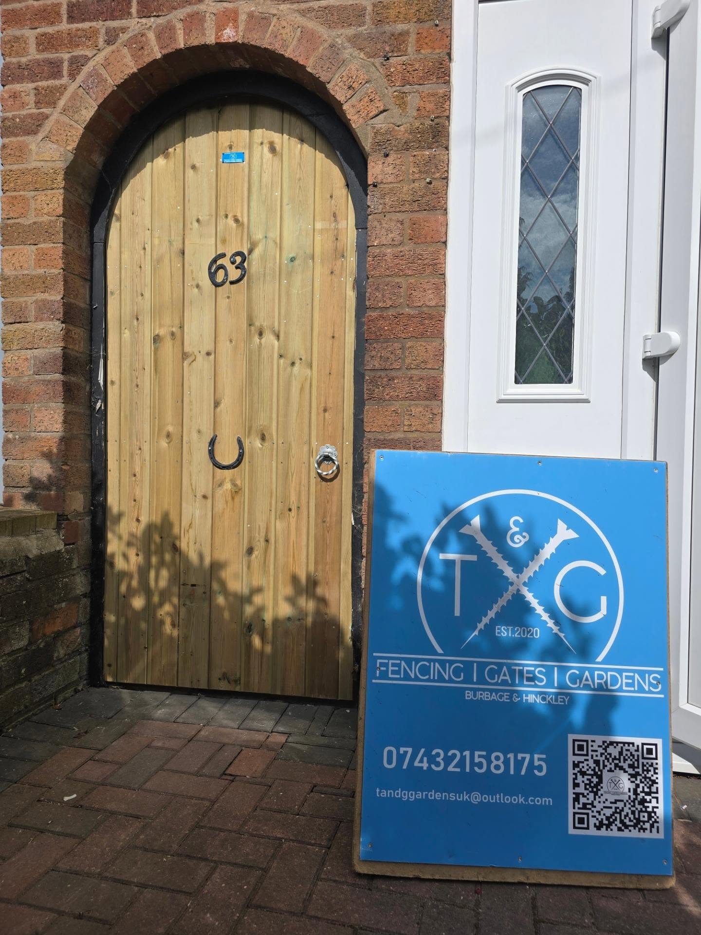 Arch-topped timber gate fitted into a brick archway beside a front door, with the T&G sign board